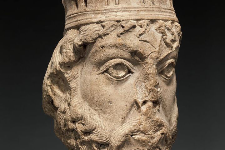 Ancient stone sculpture of a bearded man's head with a crown, against a dark background.